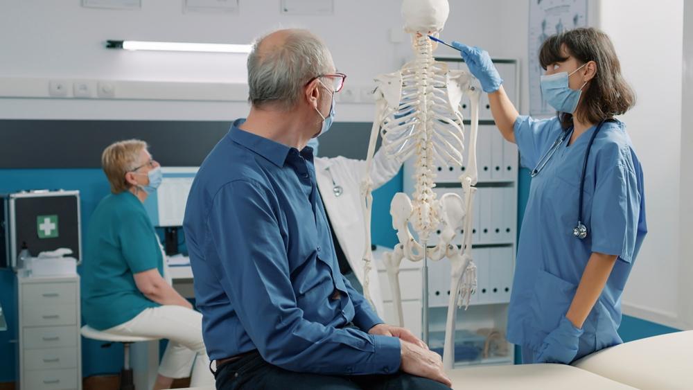 A medical specialist points to a spinal cord on a model of a human skeleton as she talks to a patient who asks about the common cause of spinal cord injuries.