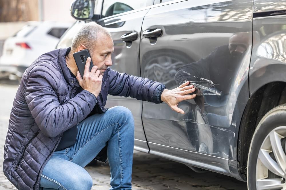 A man crouching to look at a dent in his car while he holds a phone to his ear and asks, “Can you file a car accident claim without a police report?”