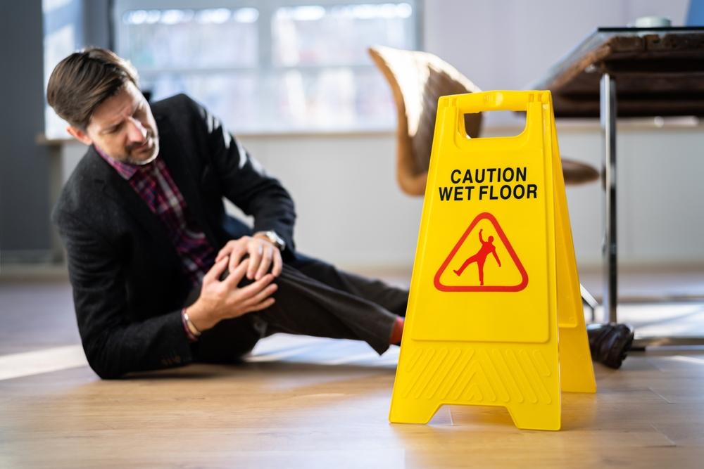 Man sits next to a wet floor sign while clutching his knee in pain and wondering who is liable in a slip and fall accident.